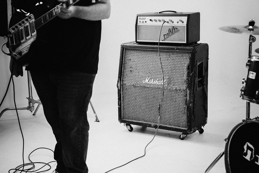 Black and white photo of a guitarist holding an electric guitar standing in front of a large amplifier