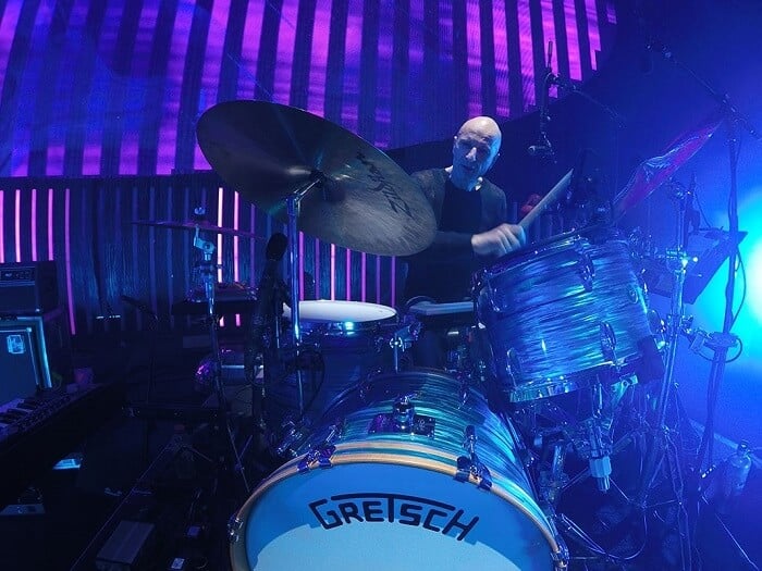 A photo of Clive Deamer playing on a Gretsch drum kit in front of purple and blue stage lighting.