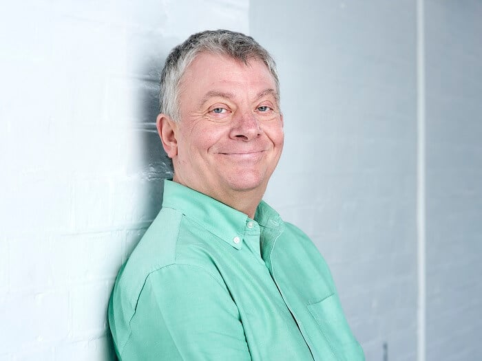 Alex Gascoine, a white man with grey hair smiles into the camera. He is leaning against a white wall and wearing a mint green shirt.