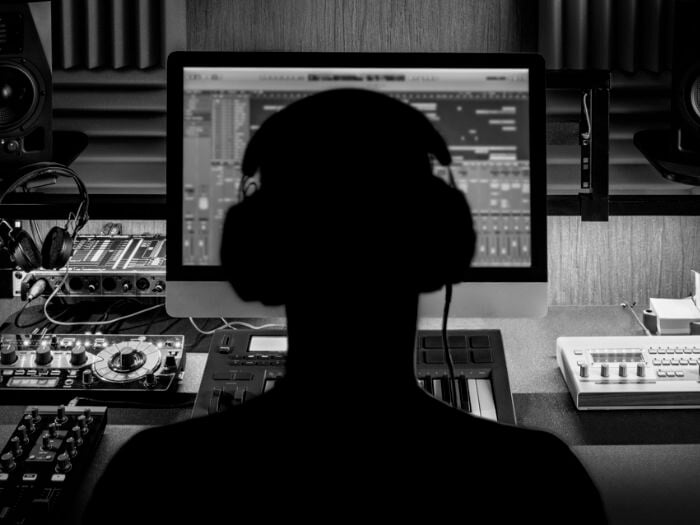 Black and white image of a musician from behind, looking at a computer screen while they compose electronic music in a studio.