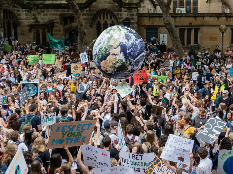 A huge crowd of climate protestrors with various banners gathered in a street, a blow up beachball style planet is being thrown by the crowd.