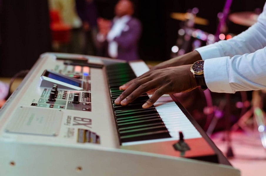 Photograph of a young persons hands on an electric piano keyboard, the background is busy and lit in purple lights - there is a blurred band playing.