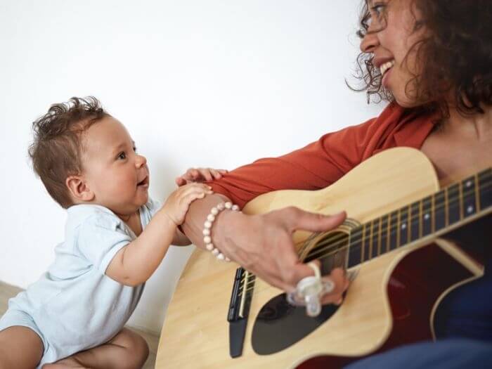 Young woman sat on the floor playing acoustic guitar next to her baby.