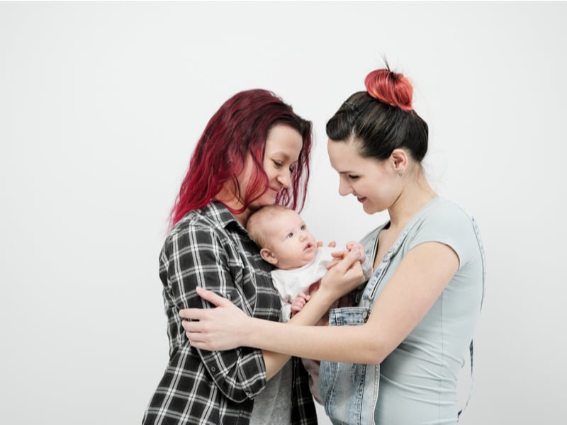Two same-sex parents embracing a new born baby. Both woman have red/pink in their hair and are looking down and smiling at the baby in the middle of them.