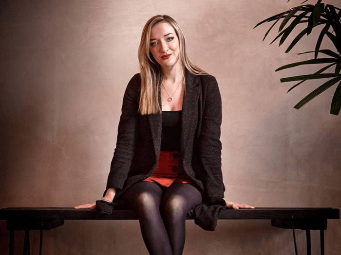 Picture of Iona Fyfe sitting on a bench in a photography studio smiling looking into the camera. She has blonde hair, red lipstick and a suit style jacket on.
