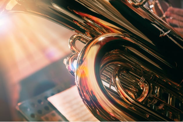 Photograph of a brass instrument being played in an orchestral context. We are close to the instrument, but can see music stands and more musicians blurred in the background.