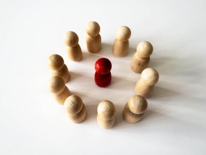 Red wooden doll figure surrounded in circle by other doll figures on a white background. Harassment and bully concept.
