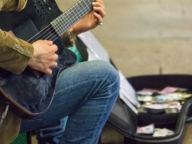 Photograph of a busker, their head isn't visible but they are wearing blue jeans and holding a plugged in electric guitar. In the background an open guitar case has money collected in it.