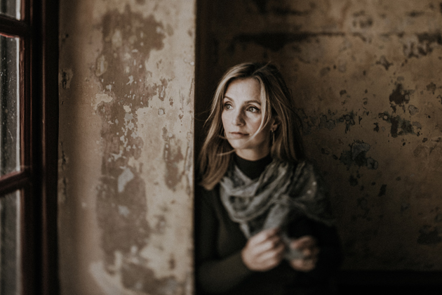 Cara Dillon looks out of a window sideways with a contemplative expression, in dim lighting. The walls behind and around her are aged and faded, suggesting that she's standing in an old cottage.