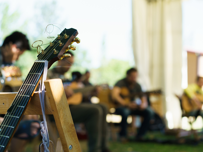 Photograph of a sunny classroom, the photo is focused on a guitar leaning against a chair, in the background out of focus you can see a number of children holding guitars.