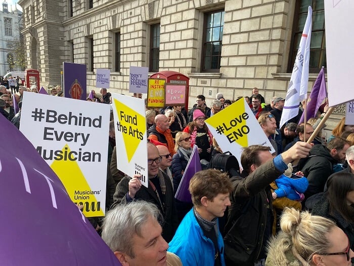 A large crowd marches in central London, some MU branded signs read Fund The Arts and Love ENO.
