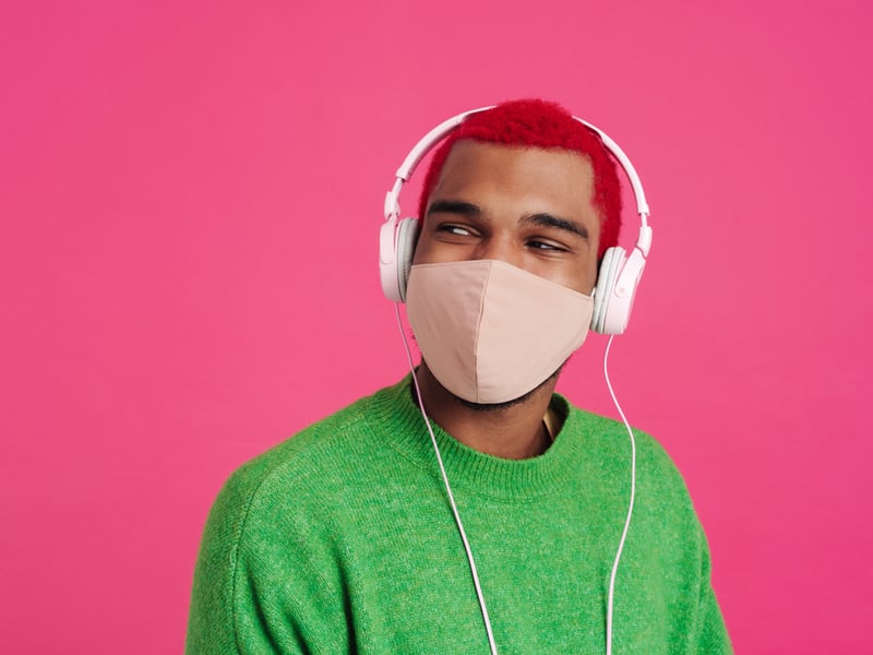 Photograph of a person with red hair, wearing a pink mask and pink earphones in front of a pink background.