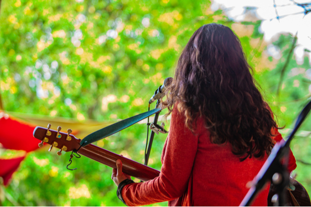 Photograph of female identifying musician playing acoustic guitar on an outdoor stage, green trees in the background