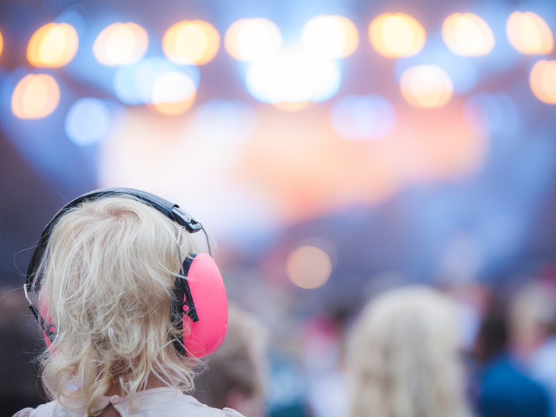 Photograph of a young girl wearing ear protectors at a concert. The background is blurred but we can see multiple people and string lights.
