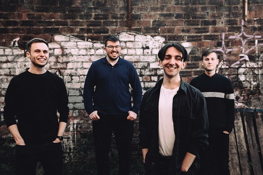 Photograph of the band Gnoss, they are four men standing in front of a brick wall. Hands in their pockets, they are smiling at the camera.