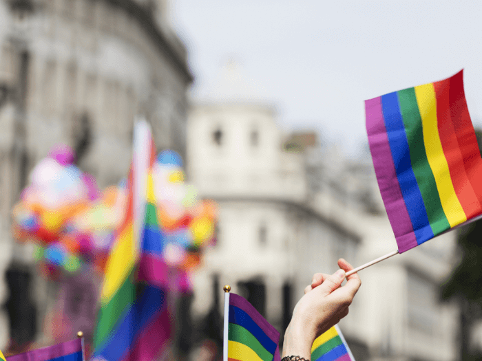 A crowd/parade with LGBT rainbow flags in the air, out of focus apart from one hand outstretched holding a flag in the foreground.