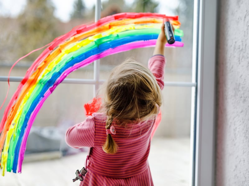 Photograph of a little kid painting a rainbow onto a big glass window.