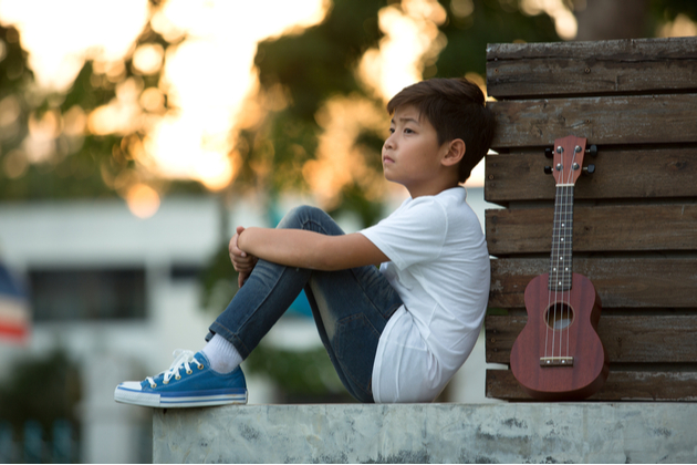 Photo of a young child, sat outdoors with a ukulele looking dreamily into the distance.