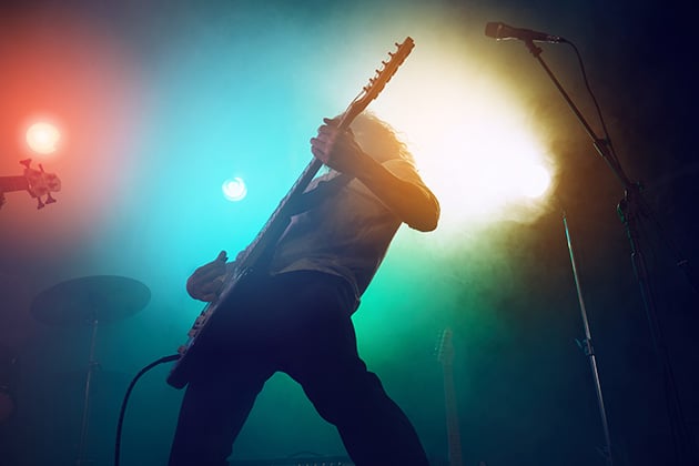 Photograph of musician playing electric guitar on stage with smokey background and lights of various colours.