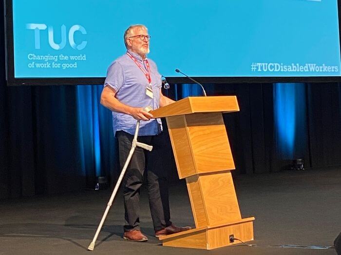 Nigel Braithwaite on stage, behind a podium, making his speech to conference holding a walking aid. Behind him is the TUC blue presentation screen with a text display of his speech.