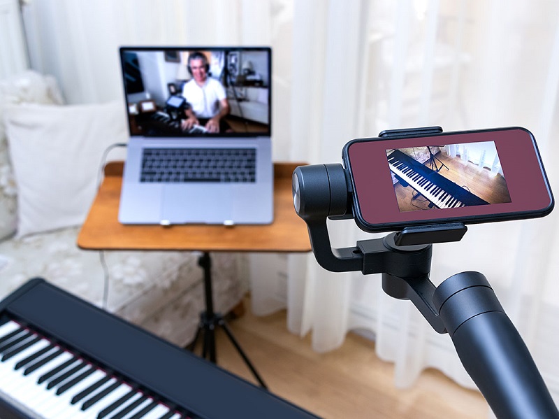 Photograph of an online teaching set up, a mobile phone camera and a laptop camera are recording a person playing on a keyboard.