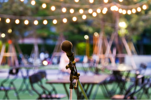 Set up for an outdoor concert, there are lights and a microphone is set up with tables and chairs in the background.