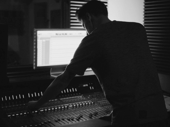 Black and white image of a session musician in the studio, looking at a screen and mixing desk.