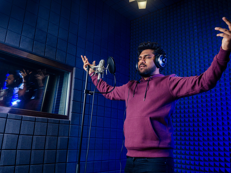 Man engrossed in singing into a microphone in a recording studio, he is dressed casually in a hooded jumper and has his arms raised and eyes closed in concentration.