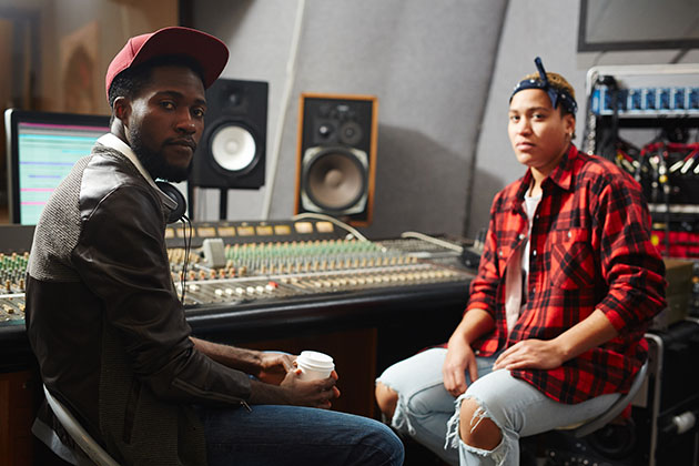 A photograph of two musicians sat in a studio next to a recording desk, they are both looking at the camera and one is holding a coffee.