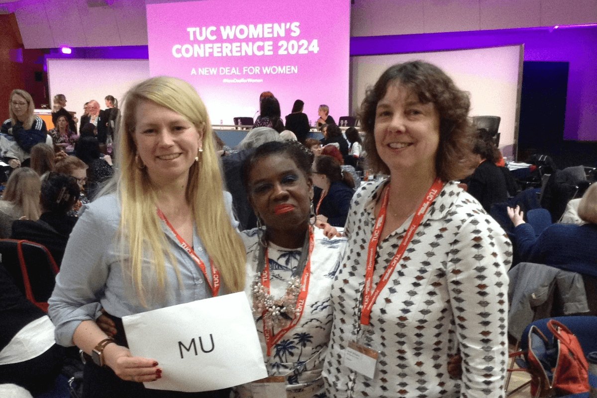 Three MU members standing at a women's conference, smiling for a photo together. One of them is holding a piece of paper saying 'MU'. In the background is a projector screen.