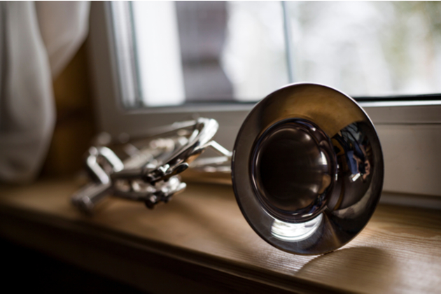 Photograph of a trumpet left resting on a window sill. The window behind the trumpet is closed.