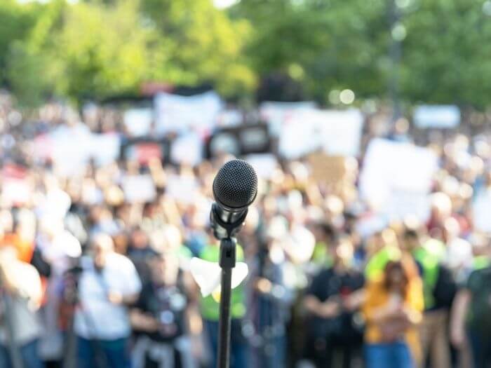 Focus on a microphone in front of a blurred crowd of people at an outdoor rally.