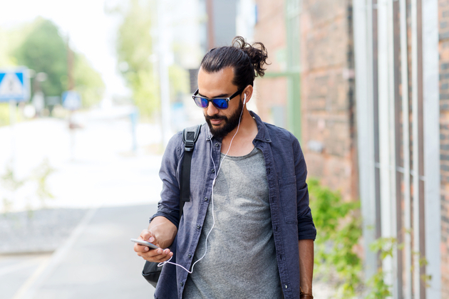 Man in sun glasses and hair tied up, wearing an open denim shirt walking down the street listening to wired headphones from phone.