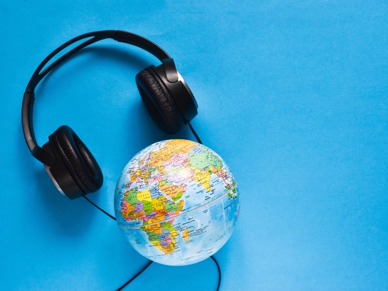 Against a blue background is a miniature toy globe that functions as a map of the world, with a large pair of black earphones sitting behind the globe.