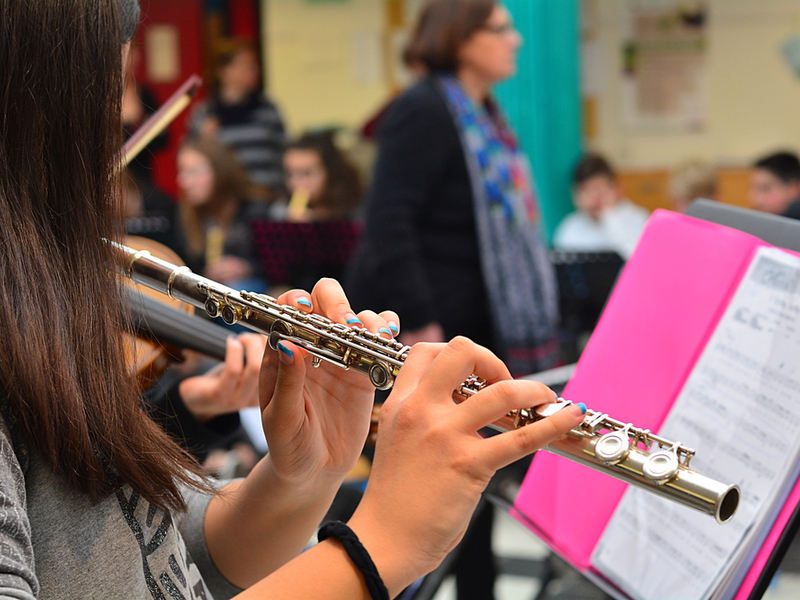 Photograph of a close up of a young flute player, practicing in a classroom setting. Behind them we can see other performers and a teacher figure.