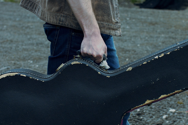 Photograph of a musician walking, whilst carrying a guitar in a hard case. We can only see the musicians arms and torso, the guitar case is well worn looking.