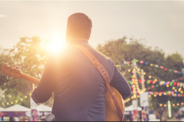 Photograph of a musician performing at an outdoor event. We can only seem them and the neck of their guitar from behind. The sun is setting and the evening lighting is turning on.