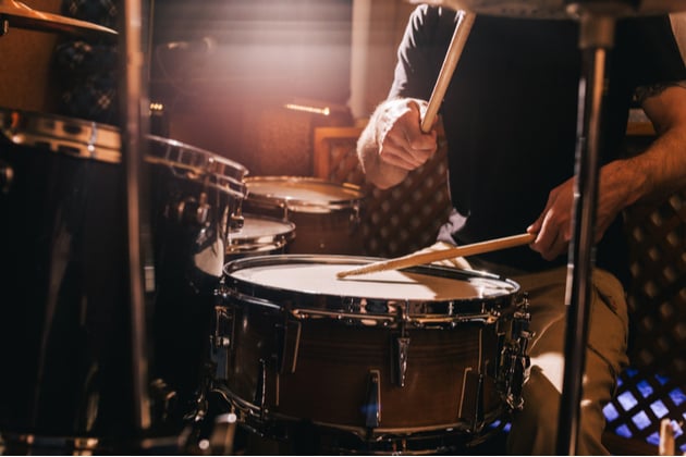 Photograph of a drum kit, a person sits behind the drum kit, we can just see their legs, arms and the drum sticks frozen in action.