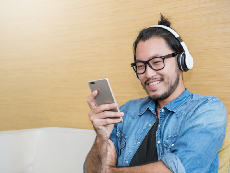 Photograph of a man sat listening to music through headphones which are connected to his mobile phone, he is smiling and happy looking.