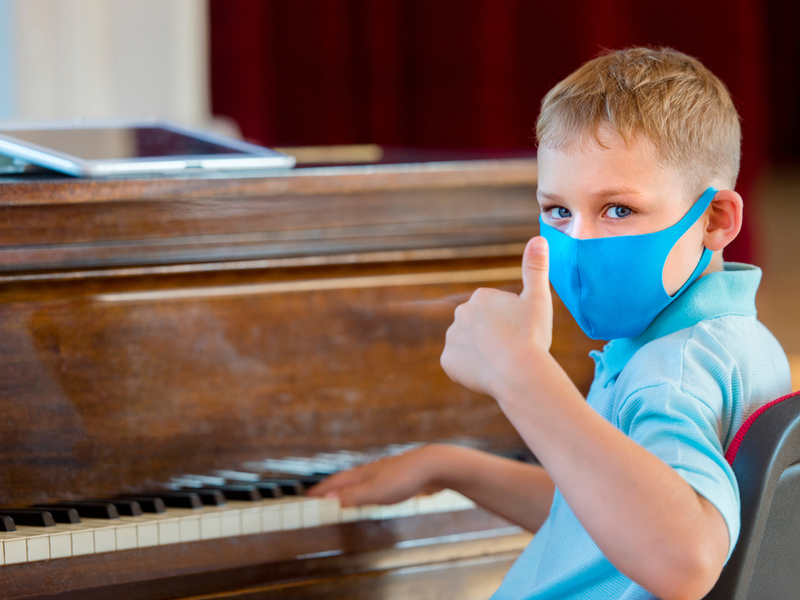 Young child wearing a mask is sat at a piano, turning round to face the camera he raises one thumb in a positive gesture.