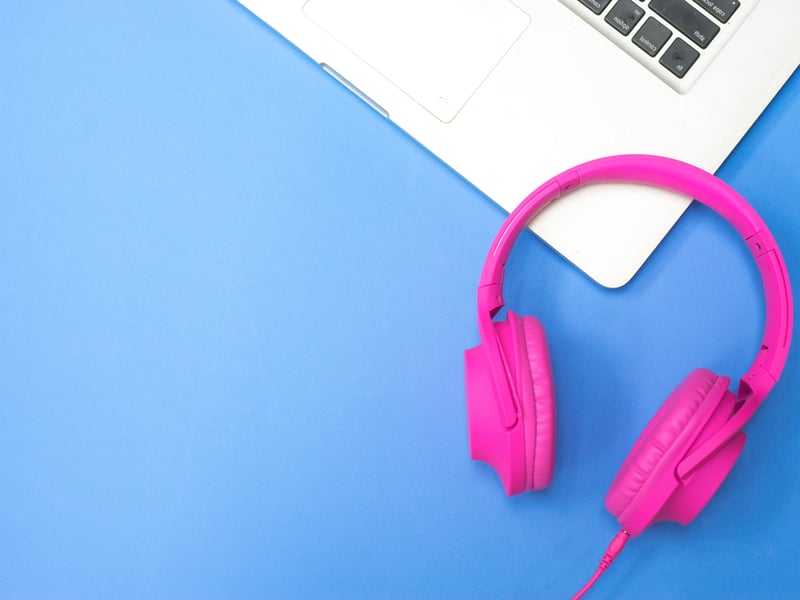 Photograph of bright pink earphones against a blue background, there is also the corner of an open silver laptop visible.