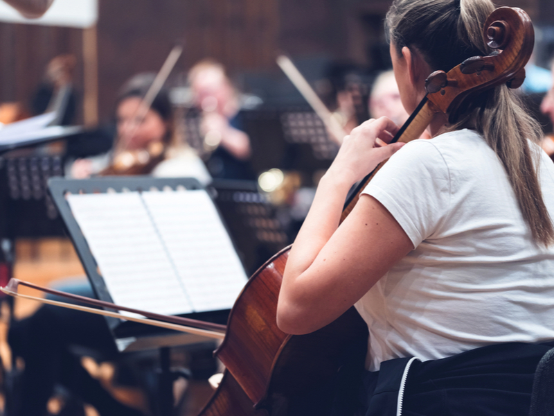 Photograph of a collection of young people rehearsing in an orchestra, the photo focuses on a young woman on the cello.