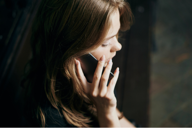 Close up of woman with light brown hair holding a mobile phone to her right ear, the back ground is dark and out of focus.